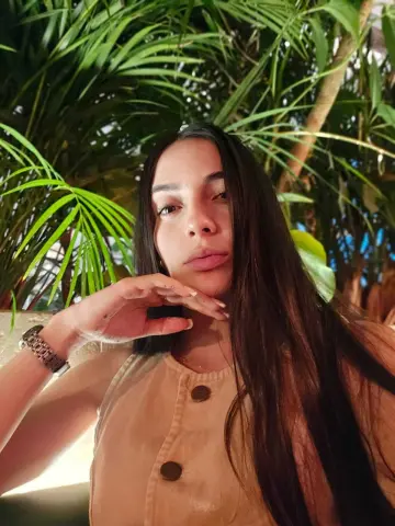 Young woman in a beige linen top posing thoughtfully amidst lush green tropical palm leaves.