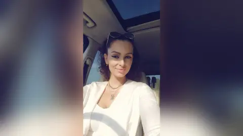 AishaApril wearing a white top and sunglasses, sitting in a car with a sunroof visible behind her.