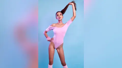 LucyMartin poses against a blue background wearing a light pink off-the-shoulder bodysuit and pink wristbands while holding her long brown hair.