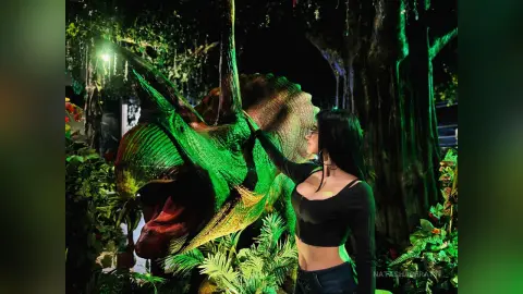Woman in a black crop top petting a large green Triceratops dinosaur statue in a lush, dark garden setting.