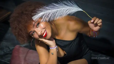 Smiling woman with afro hair and red lipstick wearing a black top, holding a white feather in a moody setting.