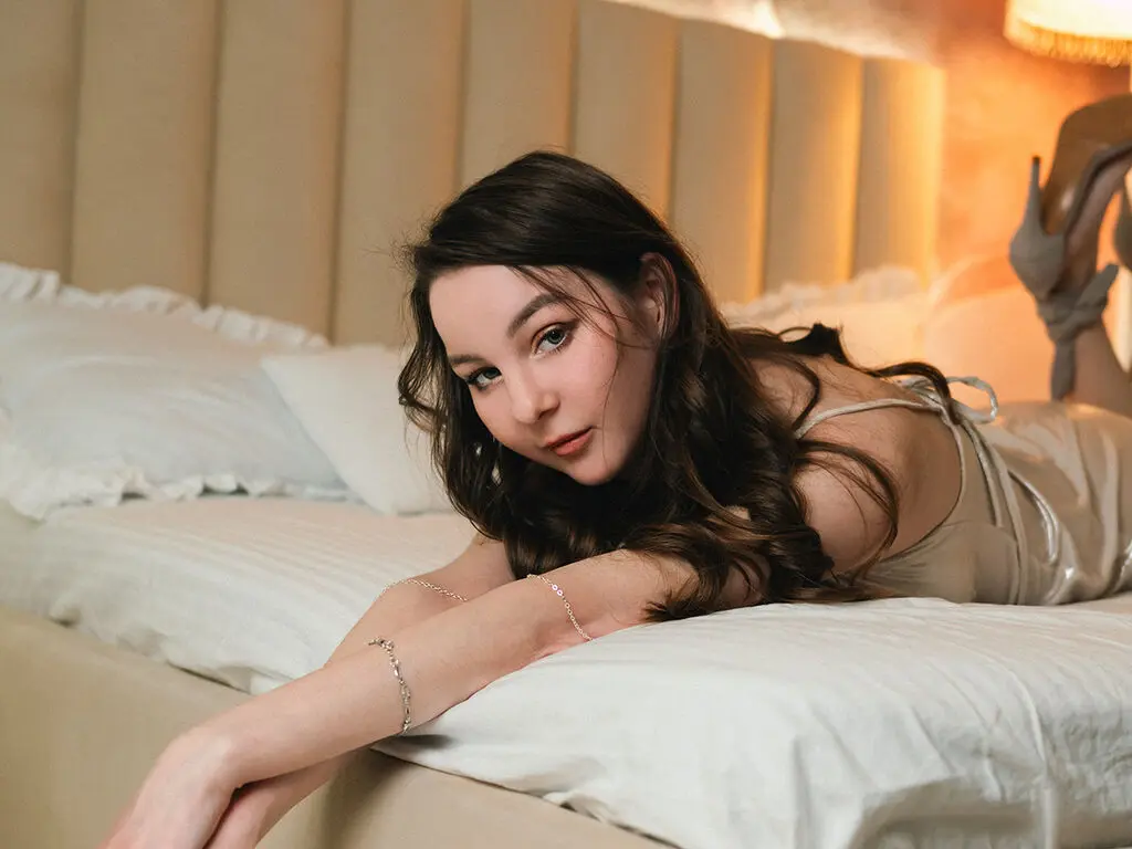 Young woman in a silk slip dress relaxing on a white bed in a warm, cozy bedroom setting.