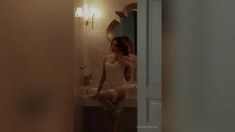Elegant woman in white lace lingerie sitting in a dimly lit, moody bathroom setting.