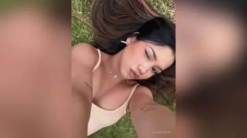 Young woman in a beige ribbed tank top relaxing on green grass for a peaceful summer outdoor selfie.