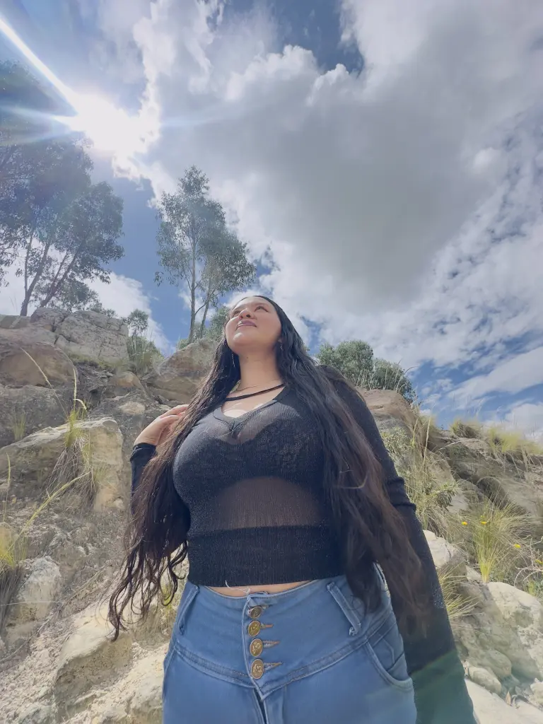 Lullabloom — Lullabloom stands outdoors against a cloudy sky wearing a black sheer long-sleeve top and blue denim jeans.