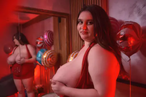 Curvy woman in a red dress posing in a festive room with red balloons and warm lighting.