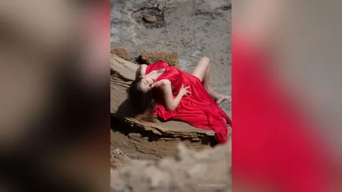 NataliaNacher wearing a long red dress while lying back on a rock outdoors.