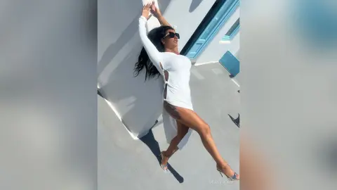 MissyJolie poses in a white long-sleeved dress and sunglasses, standing against a white wall with one leg extended.