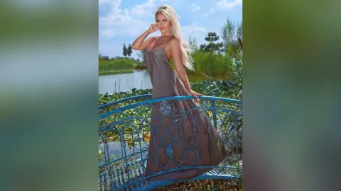 Anita stands on a blue metal balcony wearing a long, sleeveless brown dress with her blonde hair flowing down her back.