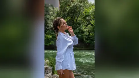 Blonde woman in a white button-down shirt posing peacefully by a lush green river in nature.