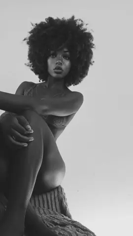 Moody black and white portrait of a woman with voluminous afro hair wearing lace lingerie in a minimalist setting.
