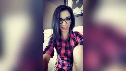Smiling woman in a red plaid shirt posing in a cozy bedroom setting.