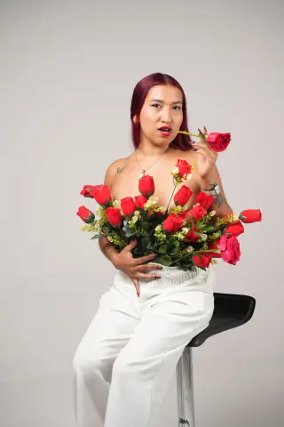 Woman in white trousers holding red roses and biting a stem against a plain background, romantic mood.