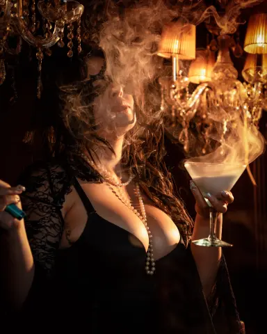 Glamorous woman in black lace outfit holding a cocktail in a moody, smoky lounge with golden chandeliers.