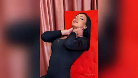 ReynaMorel poses with her eyes closed and hands behind her neck, wearing a tight black long-sleeve top against a red background.