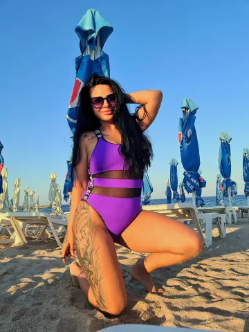 Smiling woman in a purple cutout swimsuit posing on a sunny beach under blue umbrellas.