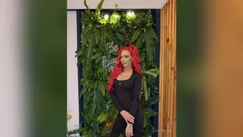 Red-haired woman in a black long-sleeve bodysuit posing against a lush green plant wall in a modern interior.