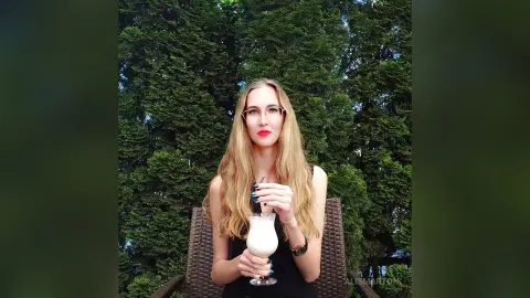 Blonde woman in a black tank top holding a milky cocktail in a lush green garden setting.