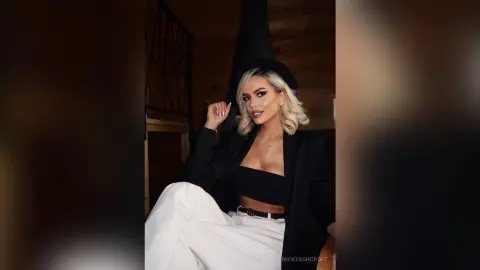 Stylish blonde woman in a black blazer and white trousers posing confidently in a moody, modern interior.