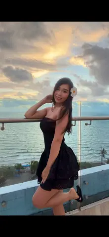 Smiling woman in a black strapless dress posing on a balcony overlooking the ocean at sunset.