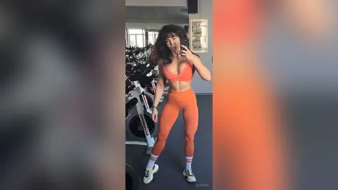 Fit woman in orange gym set taking a mirror selfie in a modern fitness center.
