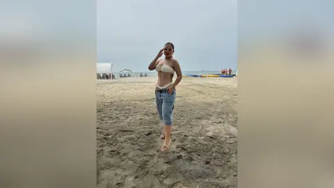 Stylish woman in a white bikini top and denim shorts standing on a sandy beach under a cloudy sky.