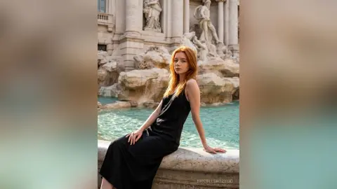 MozelleRozycki poses in a long black dress sitting on the edge of a stone fountain with her red hair down.