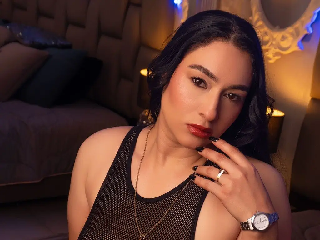 Woman in black mesh tank top and gold necklace posing in warm bedroom lighting.