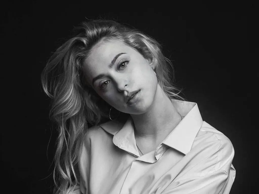 Portrait of a blonde woman in a white collared shirt against a dark background in a moody black and white style.