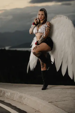 Tattooed woman in white angel wings and black outfit posing outdoors at sunset.