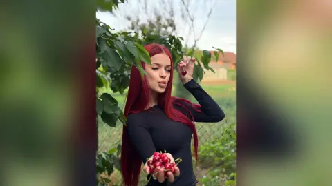 AriaVex poses outdoors with long red hair, wearing a black long-sleeve top and holding red cherries.