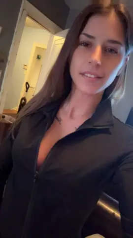 Smiling brunette woman in a black zip-up athletic jacket relaxing in a cozy indoor setting.