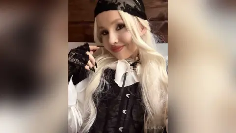EllieFrosty poses with long blonde hair and black lace headwear, wearing a black lace and white ruff-collared outfit.