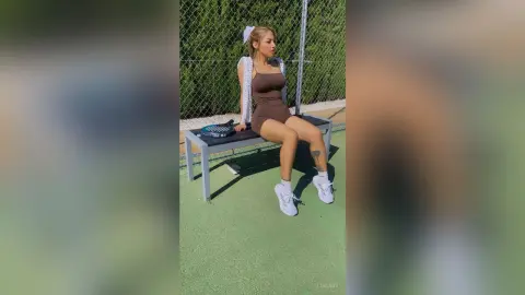 Young woman in brown athletic outfit sitting on a bench at an outdoor tennis court on a sunny day.