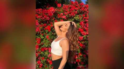 InesMoore poses with her hands behind her head, wearing a white crop top and black bottoms against a wall of red roses.