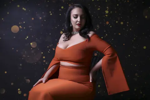 Elegant woman in an orange off-the-shoulder gown posing against a dark sparkling bokeh background.