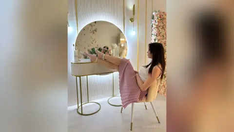 Woman in white top and pink pleated skirt relaxing in a bright, modern luxury dressing room with gold accents.