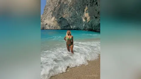 Anita stands in the shallow turquoise ocean waves wearing a green bikini on a sandy beach near a large cliff.
