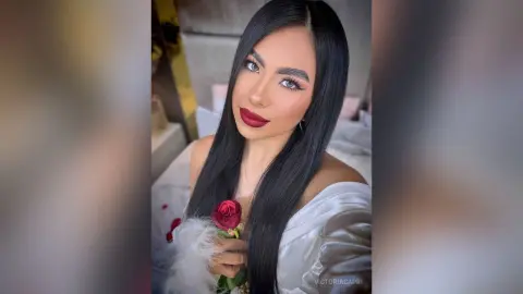 Glamorous woman with long black hair and red lipstick holding a red rose in a soft, romantic setting.