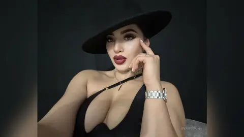 Glamorous woman in a black dress and wide-brimmed hat wearing gold jewelry in a moody studio setting.