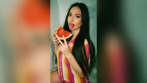 Young woman in a colorful crochet top eating watermelon against a light backdrop with a playful mood.