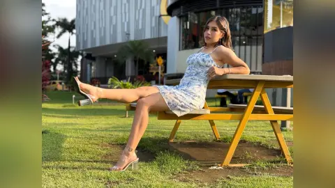 Stylish woman in a white floral mini dress sitting on a wooden picnic table in a sunny outdoor park.