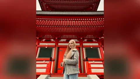 AletaKunde poses in front of a red traditional building, wearing a grey jacket and light blue jeans with her long blonde hair down.