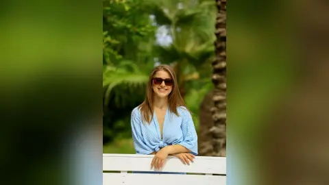 Smiling young woman in a blue linen shirt relaxing in a lush tropical garden setting.