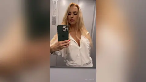 Blonde woman in a white zip-up hoodie taking a moody mirror selfie in a modern bathroom.