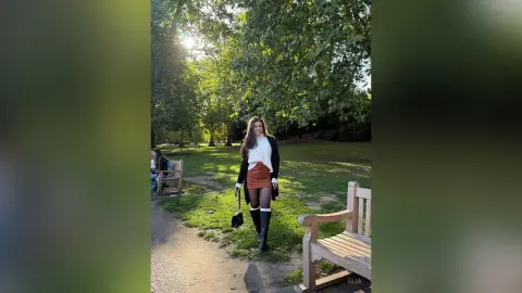 Smiling woman in a white sweater, brown skirt, and black boots walking through a sunny green park.