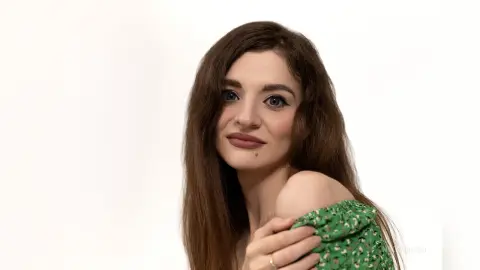 Smiling young woman with long brown hair wearing a green floral print top against a white background.