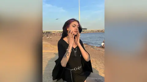 LorenLoiss poses outdoors on a sandy beach wearing a black top and silver chain belt, touching her face with her hands.