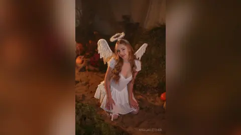 StellaFletcher wears a white lace dress and white wings while kneeling on sand in a garden setting.