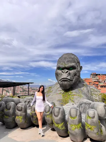 Woman in a white mini dress standing next to a massive stone gorilla statue under a cloudy sky.
